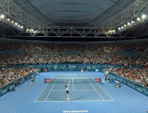 A tennis match at the Brisbane International. The tennis court is blue and a large crowd of people are watching the men's singles game..