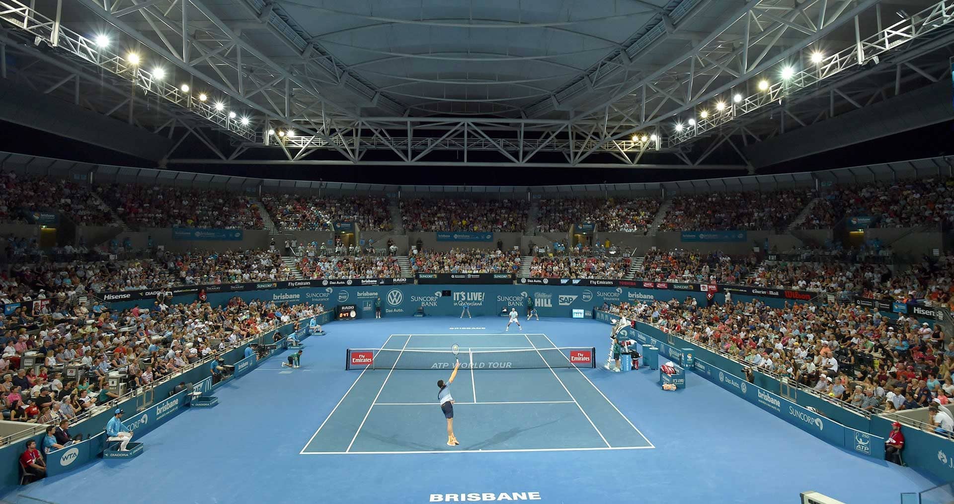 A tennis match at the Brisbane International. The tennis court is blue and a large crowd of people are watching the men's singles game.. 