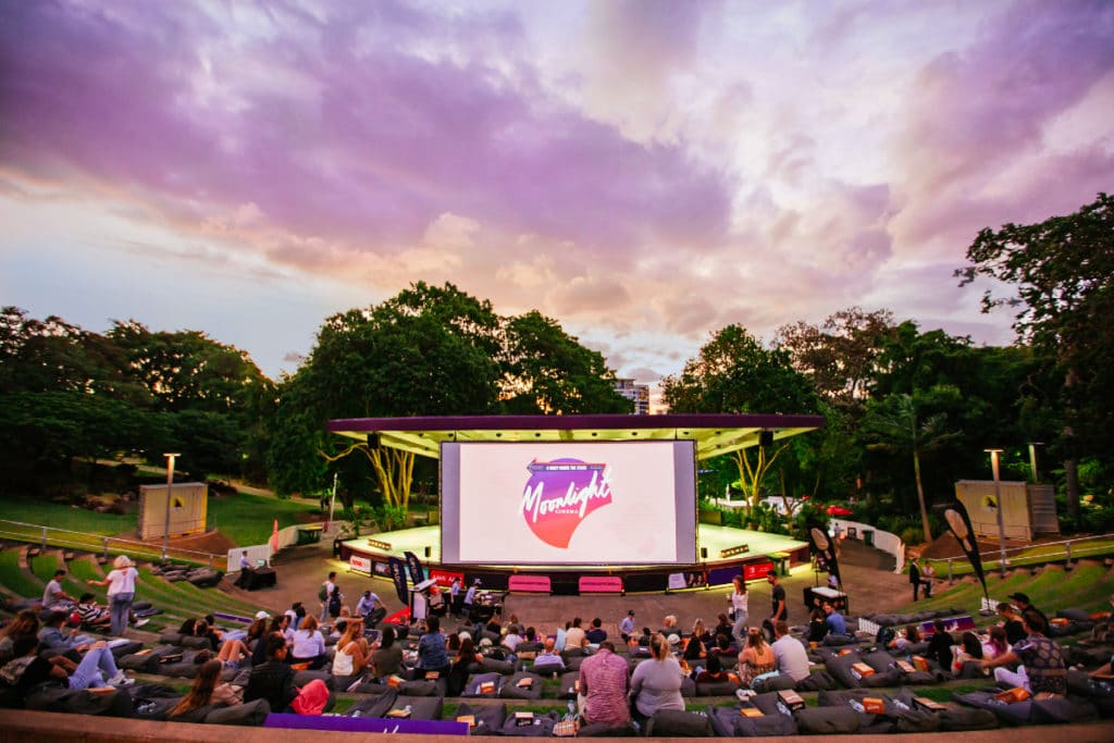 A large outdoor cinema screen with a large crowd of people on beanbags watching the movie.